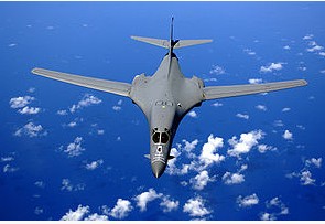 Top view of B-1B in-flight with white clouds scattered underneath. Its wings are swept fully forward as the gray aircraft flies over the ocean.
